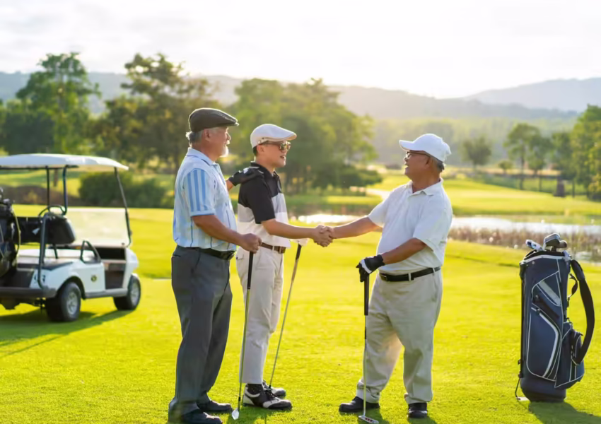 Three golfers exchanging handshakes on pristine fairway with golf cart and water feature, backdropped by African landscape at sunrise.