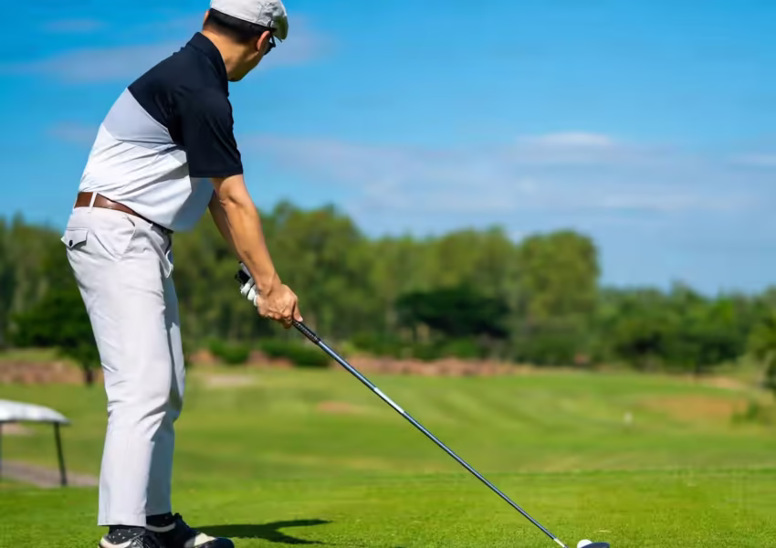 Golfer in classic white and black attire positioning for a drive shot on a pristine championship course bordered by native African woodland under clear blue skies.