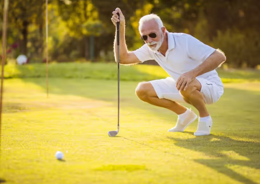 Senior golfer in crisp white attire analyzing his putt on a sunlit green, wearing aviator sunglasses and displaying perfect form at an exclusive golf course